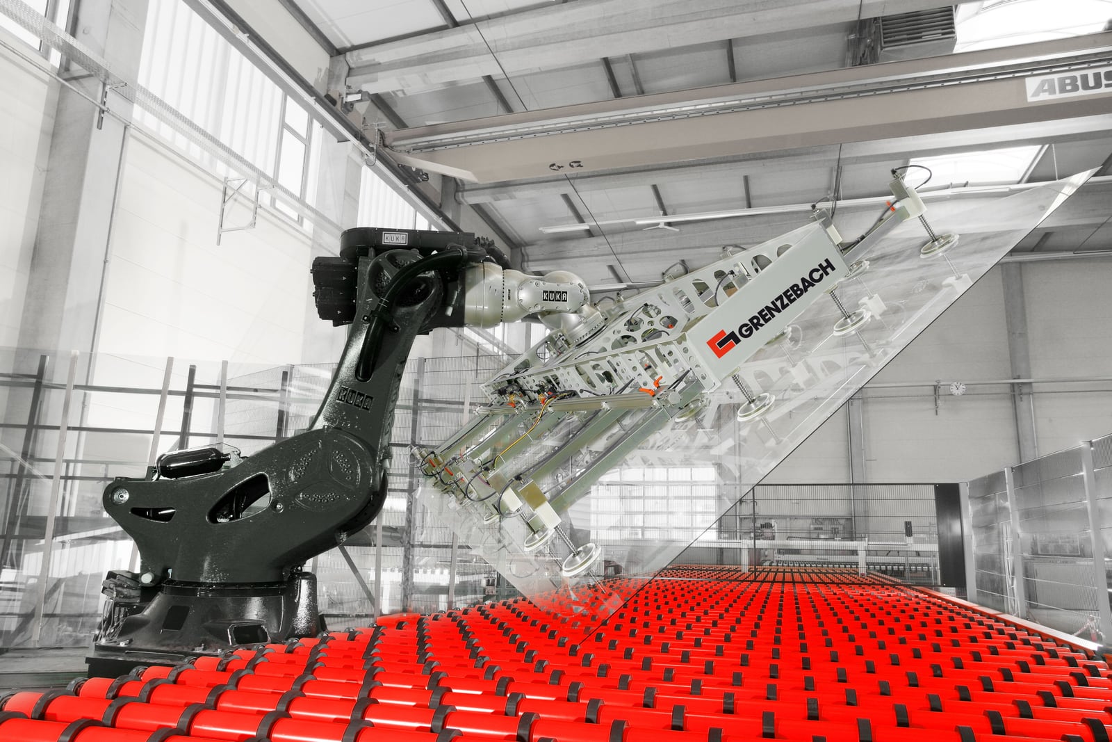 Industrial robot lifting a large glass sheet above a roller conveyor in a manufacturing plant.